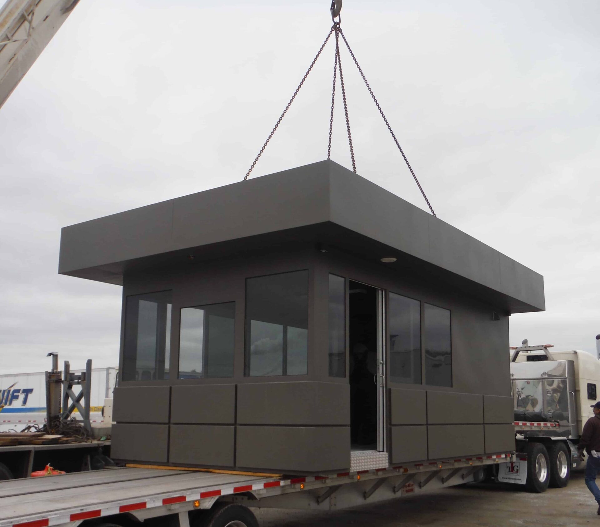 A prefabricated guard shack being lifted by a crane from a flatbed trailer at an industrial site.