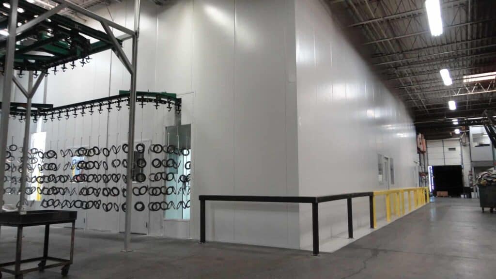 A warehouse interior with shiny metal car parts hanging on a rack, a table to the left side, and a white and yellow railing running parallel to clean, tall walls.