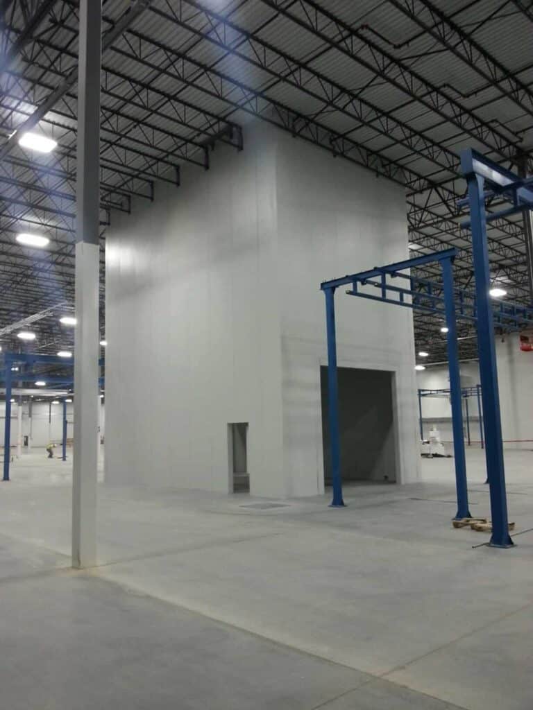 A large, empty industrial warehouse with tall walls, featuring structural columns painted blue, a small office space built within, and expansive gray floors.